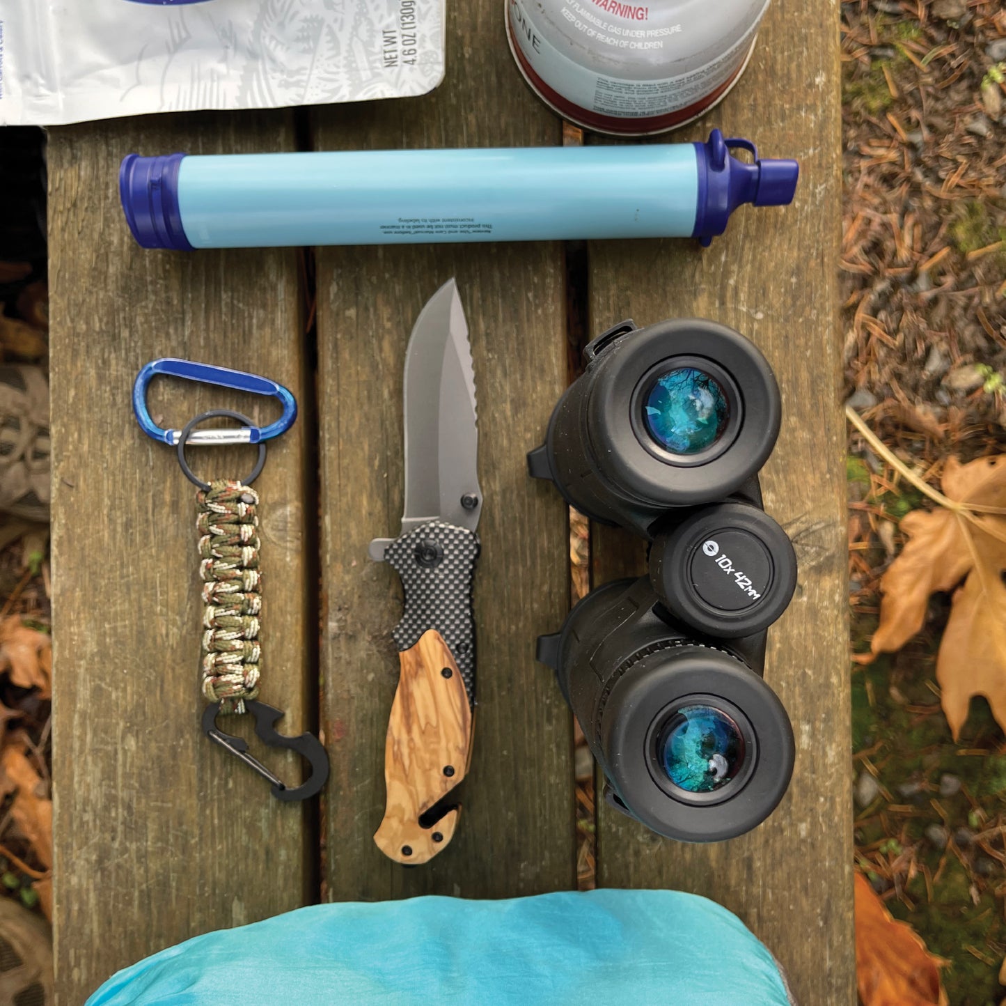 Knife, binoculars, and other outdoor gear on a wooden surface with leaves in the background
