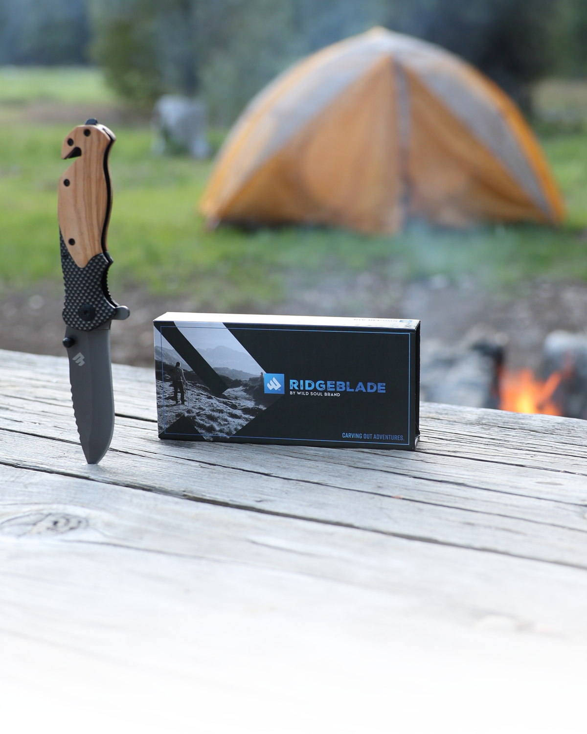 Ridgeblade folding pocket knife and its gift box on a picnic table at a campsite, with a tent and campfire behind