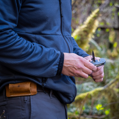 Man in the woods sharpening a folding knife on an Edge Sherpa keychain knife sharpener, leather holster on his belt