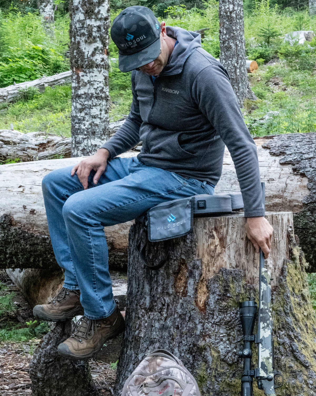 Hunter in a Wild Soul cap sitting on a tree stump in the woods on a NextPad Camp heated seat cushion, rifle beside him
