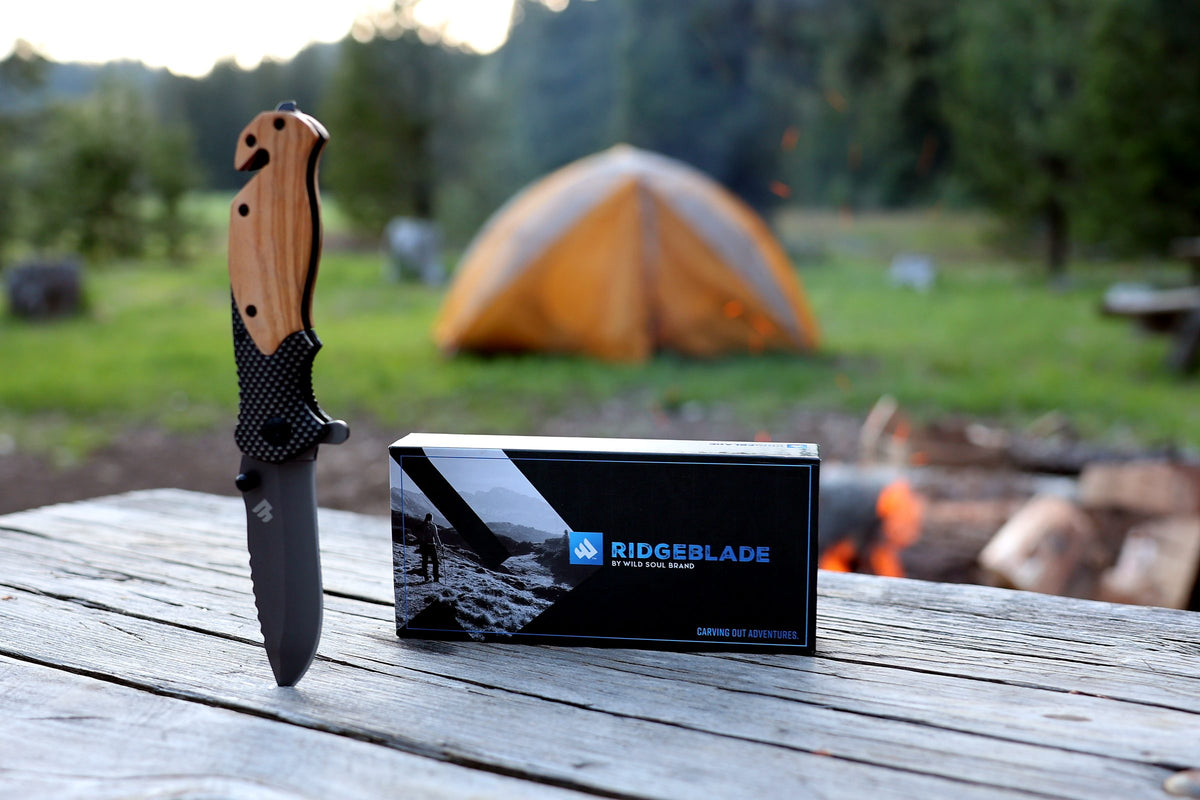Ridgeblade folding pocket knife and its gift box on a picnic table at a campsite, with a tent and campfire behind