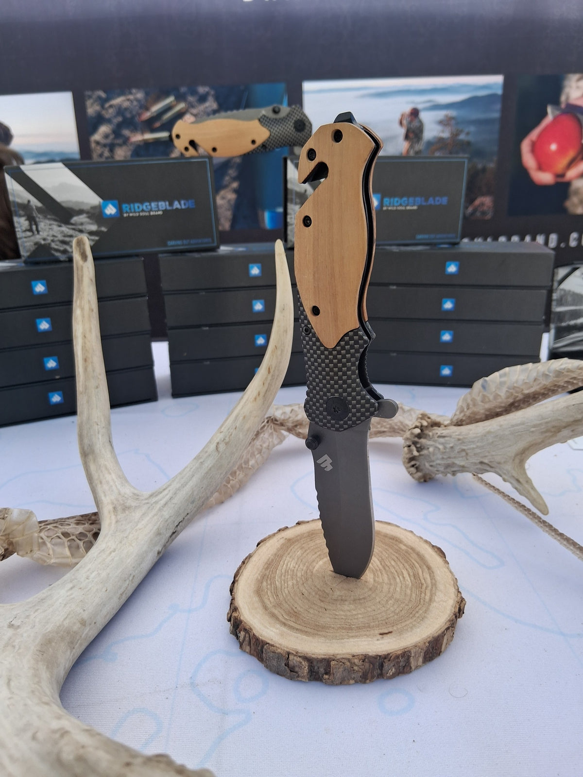 Ridgeblade folding knife displayed on a wood slice between deer antlers at an event booth