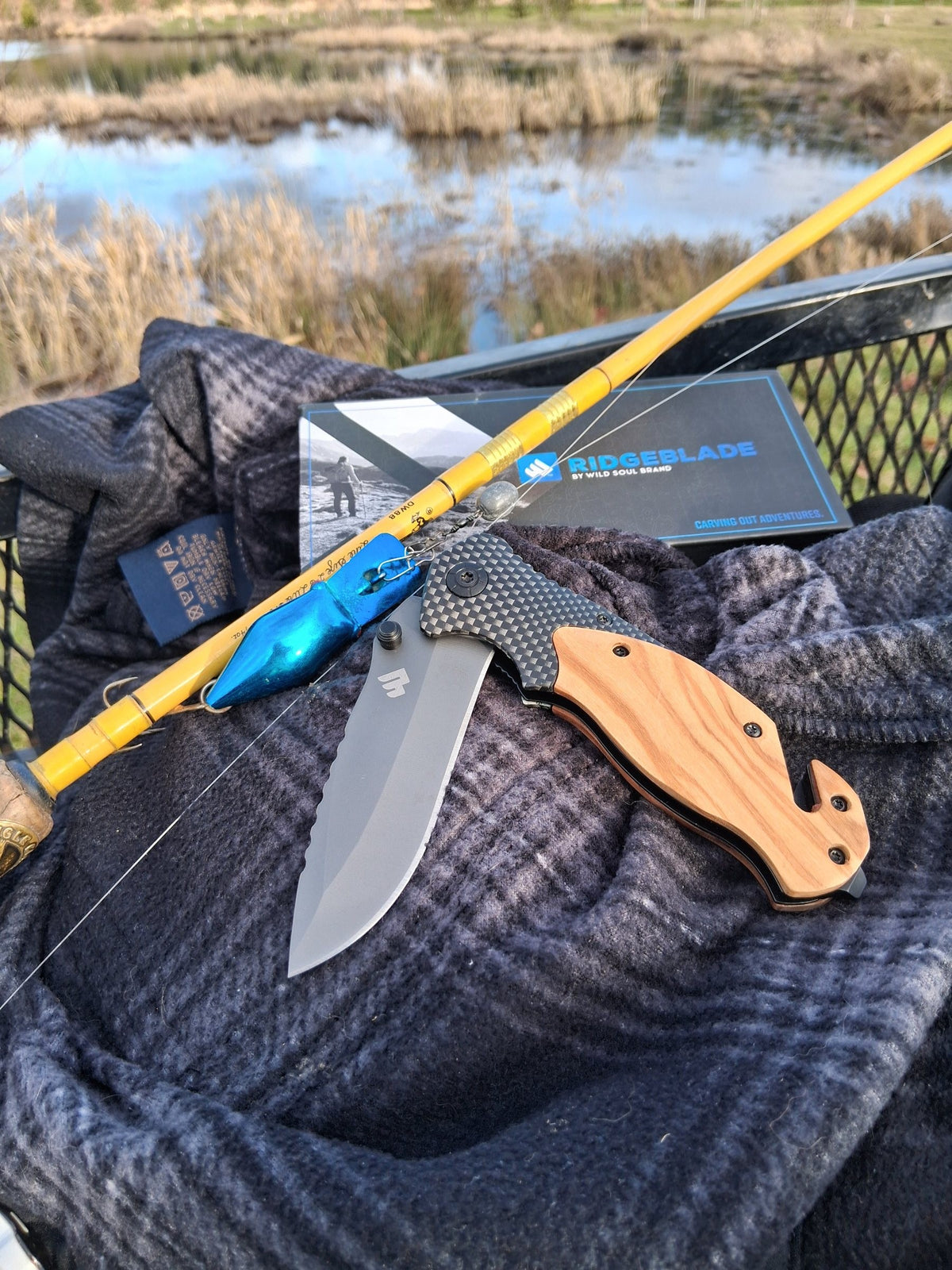 Ridgeblade folding pocket knife open on a plaid blanket beside a fishing rod at a marsh
