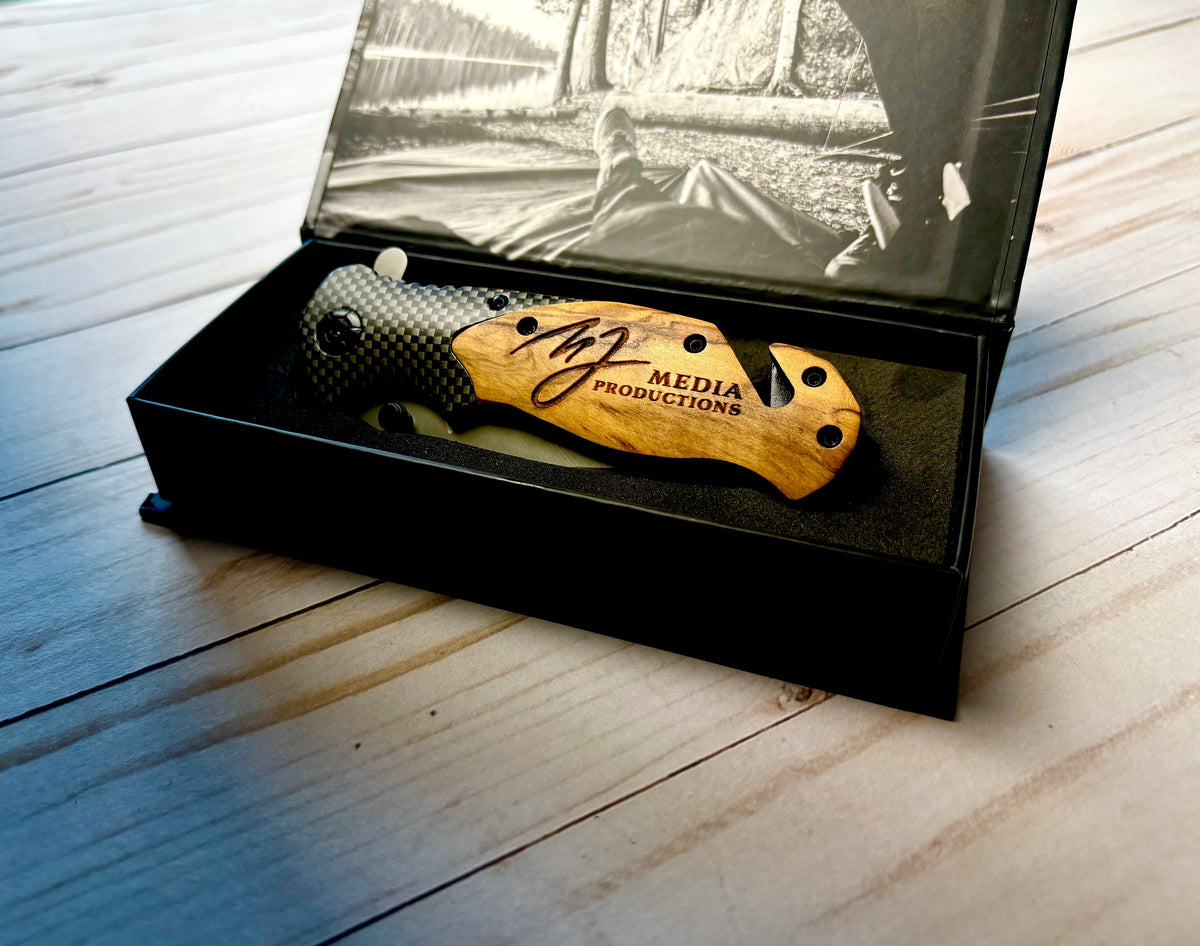 Ridgeblade folding knife laser engraved with the MJ Media Productions logo, resting in its open Wild Soul Brand gift box on a wood table