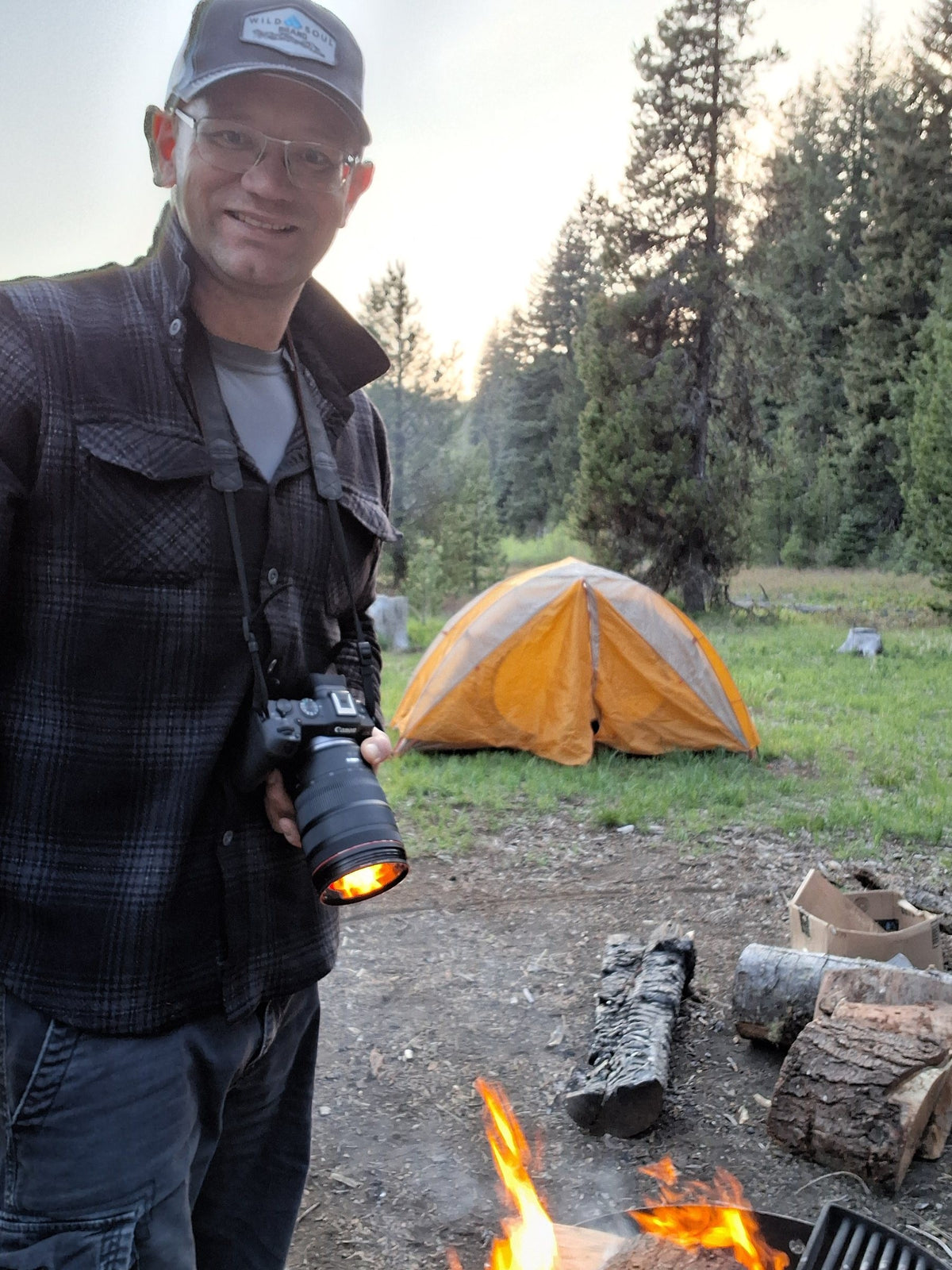 Founder with a camera beside a campfire and an orange tent in a mountain meadow