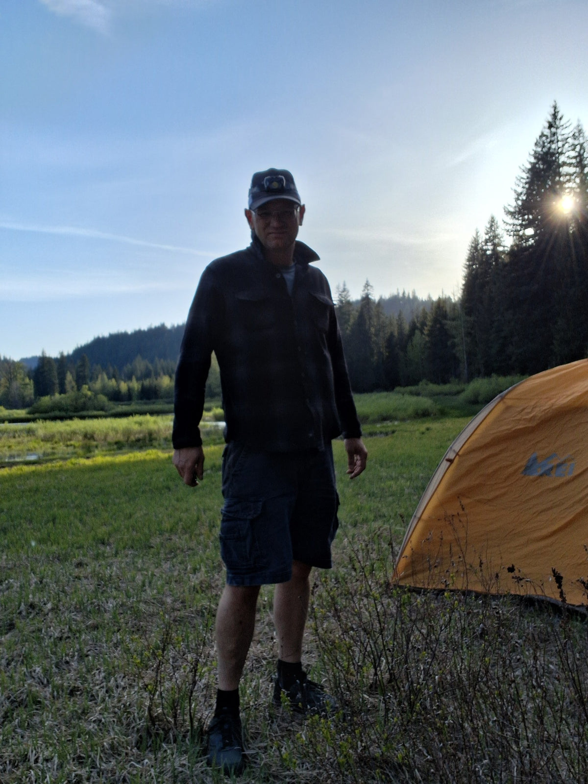 Founder standing beside an orange tent in a mountain meadow at sunset