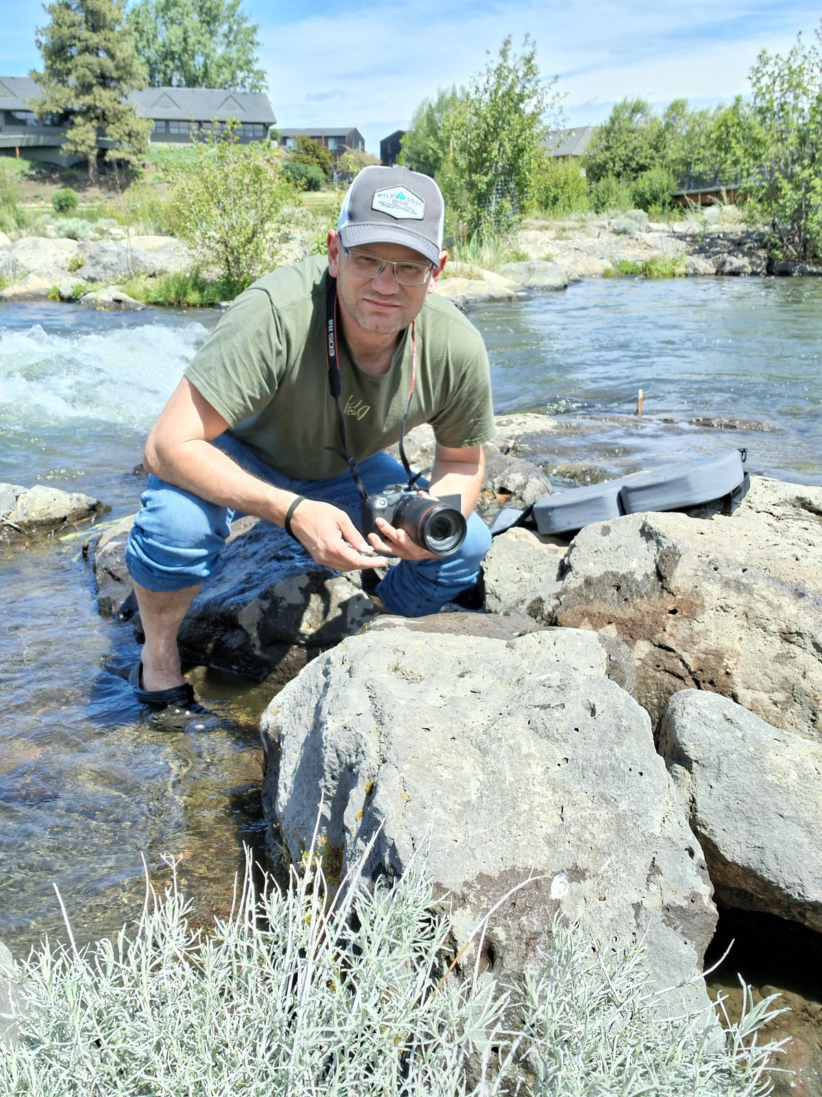 Founder crouched on river rocks holding a camera